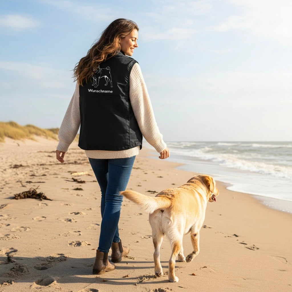 Frau in Weste mit Hundemotiv geht mit Labrador am Strand spazieren
