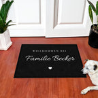 Fußmatte „Willkommen bei Familie Becker“ in Schreibschrift mit kleinem Herz, vor einer weißen Haustür, daneben liegt ein Hund