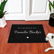 Fußmatte „Willkommen bei Familie Becker“ in Schreibschrift mit kleinem Herz, vor einer weißen Haustür, daneben liegt ein Hund