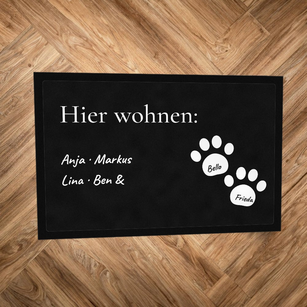 Fußmatte „Hier wohnen“ mit Familiennamen und zwei weißen Pfoten mit Hundenamen, auf Parkettboden