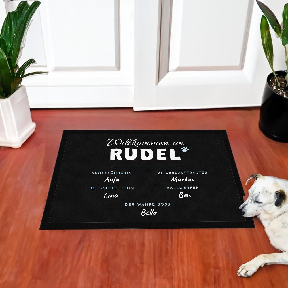 Fußmatte „Willkommen im Rudel“ mit Rollen wie Rudelführerin und Der wahre Boss und Namen, vor einer weißen Haustür, daneben liegt ein Hund