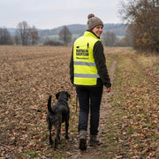 Frau mit Hund traegt Softshell Hundesportweste Mantrailing Edition neongelb auf Feldweg
