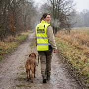 Frau mit Hund traegt Softshell Hundesportweste Mantrailing Edition neongelb auf Waldweg